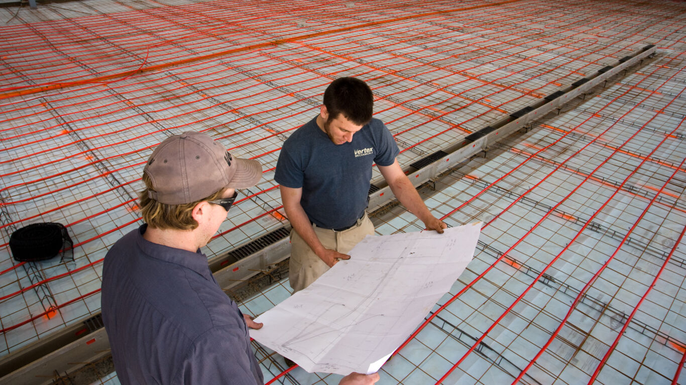 Two men looking at planning documents with radiant heating piping installed behind them on the floor.